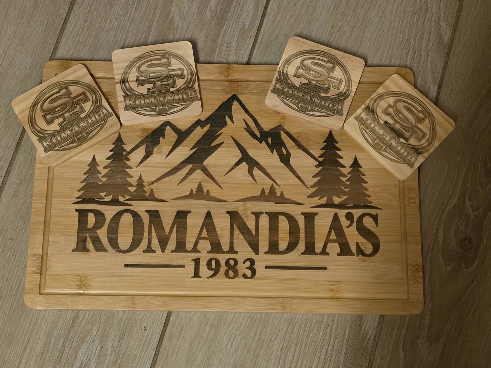 Engraved cutting board and four coasters reading ROMANDIA'S 1983 with a mountain scene